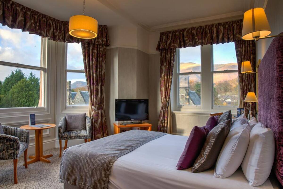 A hotel room with a large bed, two armchairs, a small table, a TV, and curtains.
