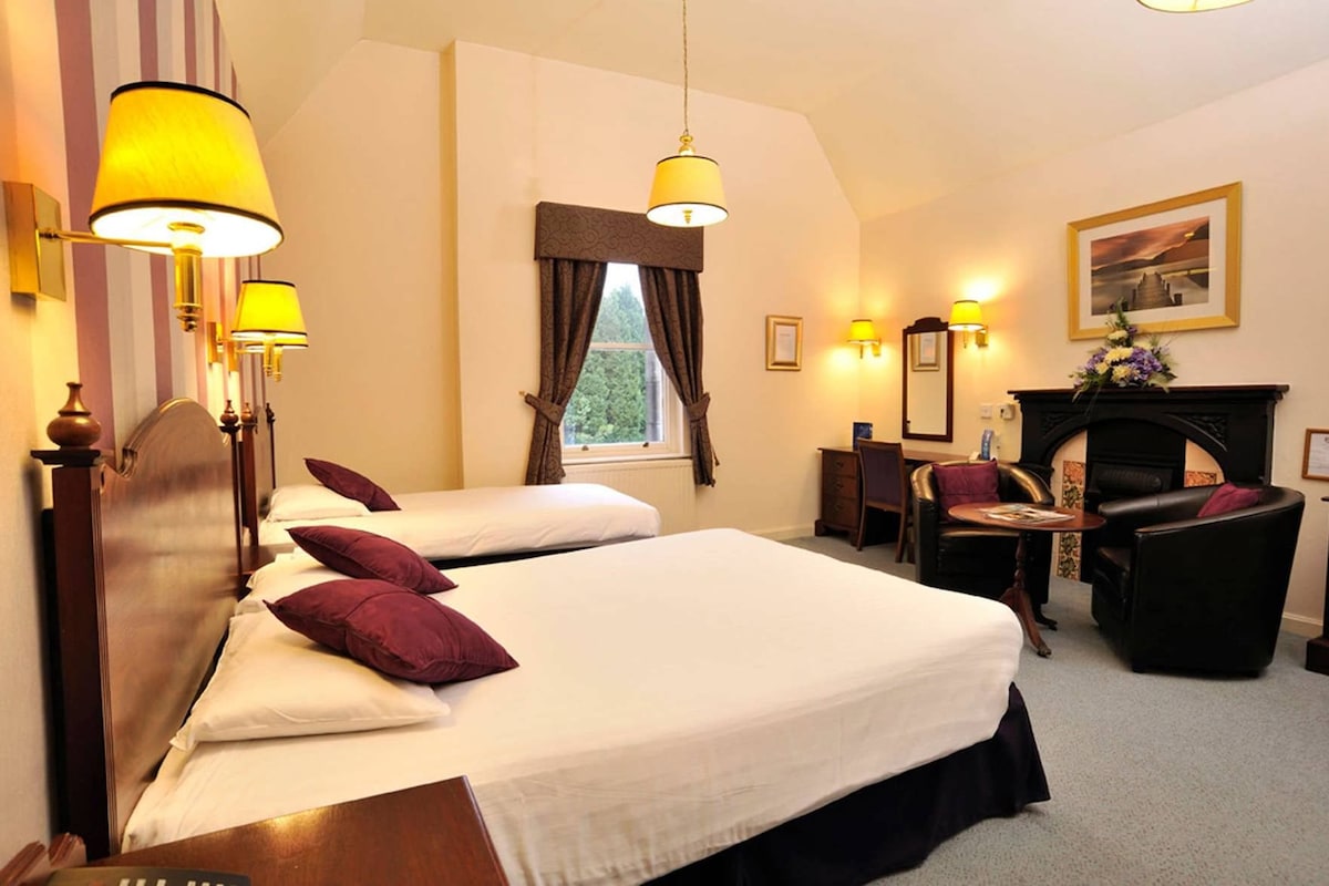 A hotel room with two beds, a desk, a chair, a fireplace, and a window with curtains.