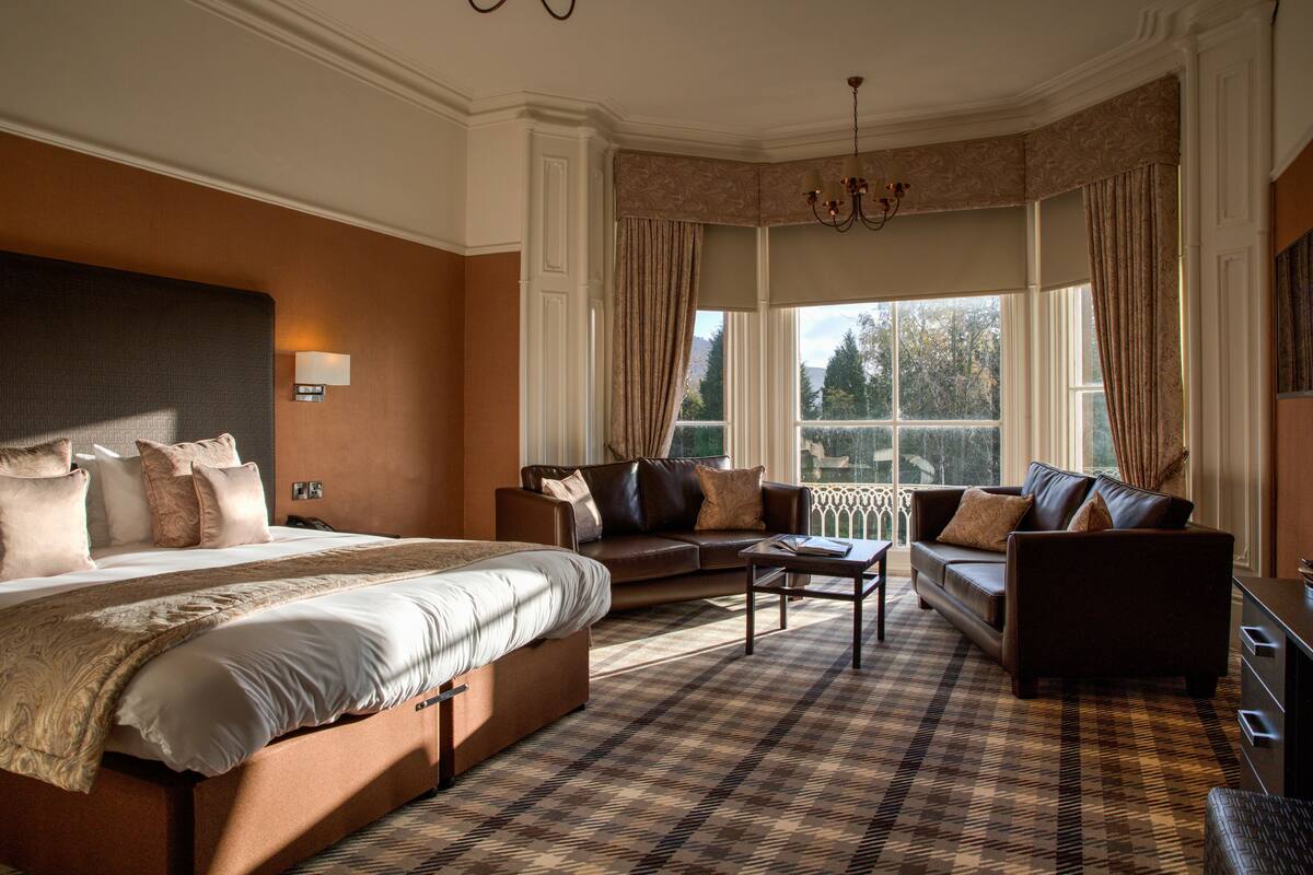 A spacious hotel room with a large bed, two sofas, a coffee table, and a view of the outdoors through large windows.