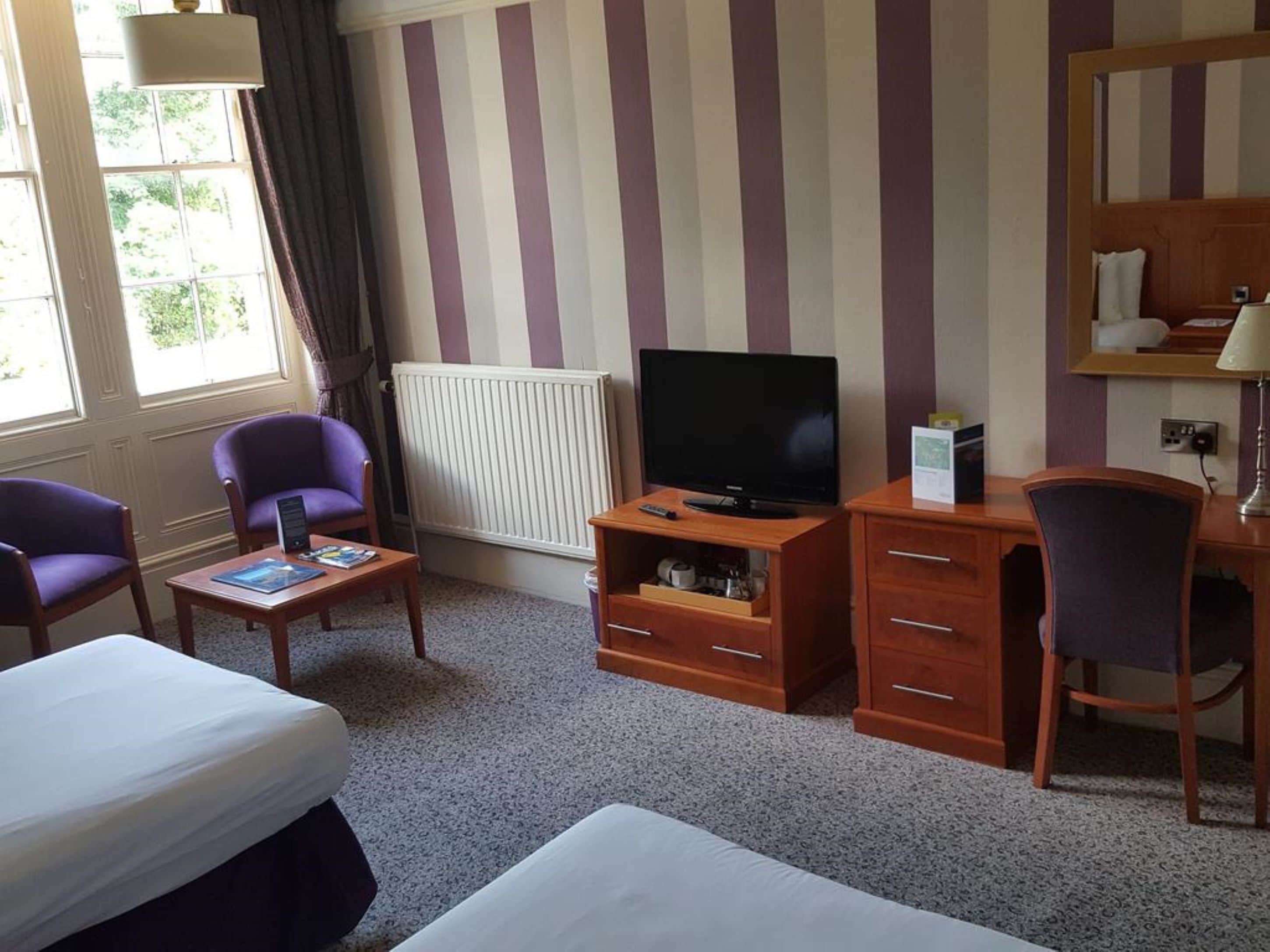 A hotel room with striped wallpaper, a bed, a desk with a TV, two purple chairs, and a radiator.