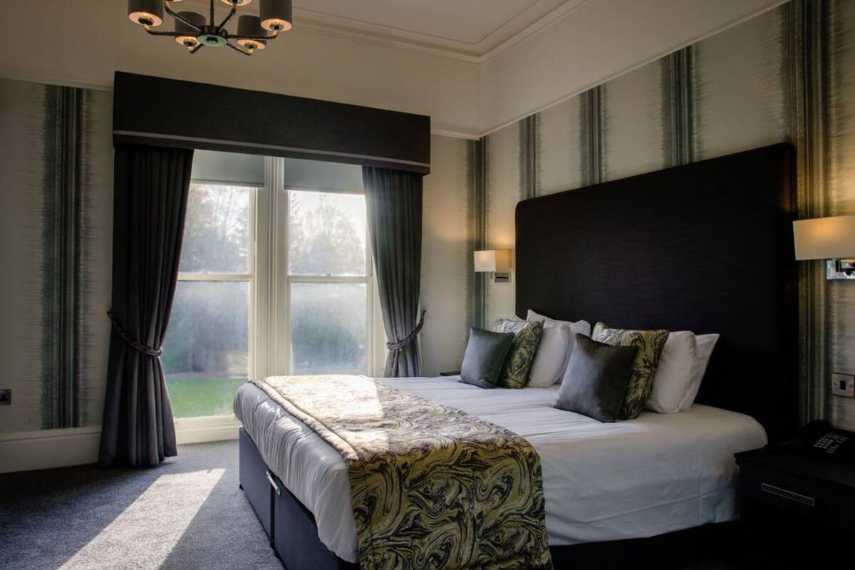 A bedroom with a large bed, a headboard, a bedside table, a lamp, and a window with curtains.