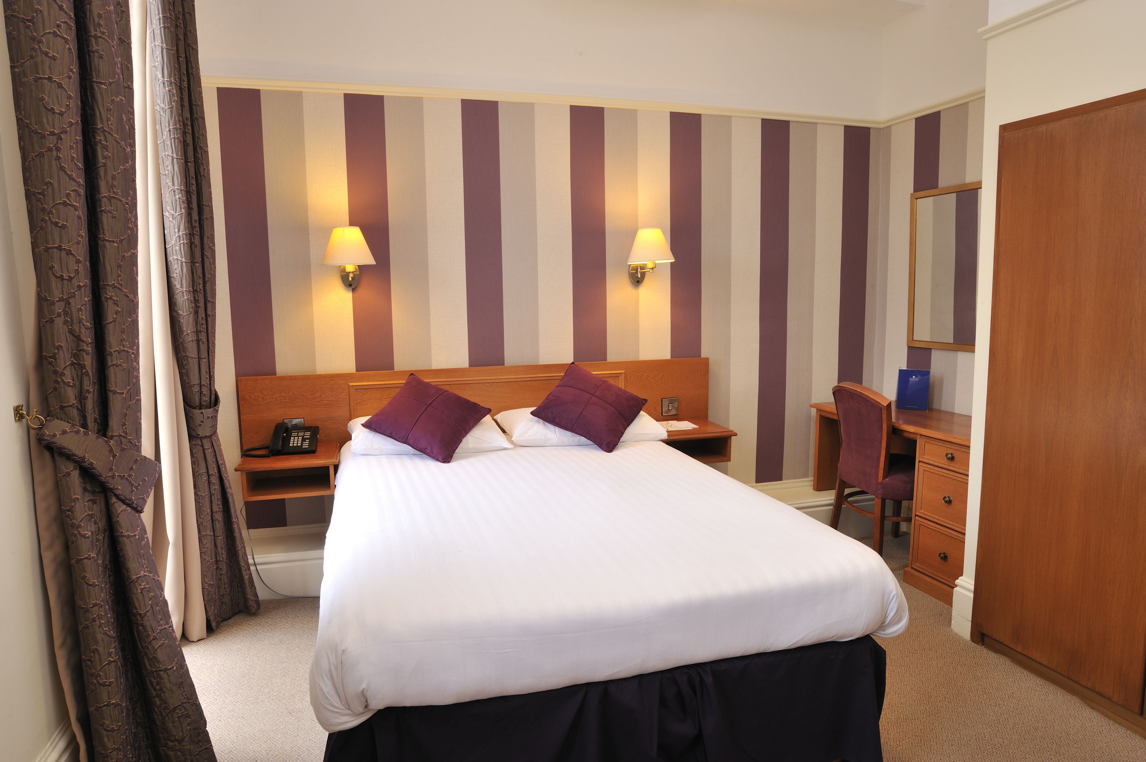 A hotel room with a large bed, two bedside lamps, a desk with a chair, a wardrobe, and a mirror.