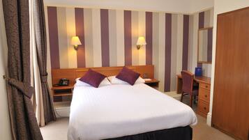 A hotel room with a large bed, two bedside lamps, a desk with a chair, a wardrobe, and a mirror.