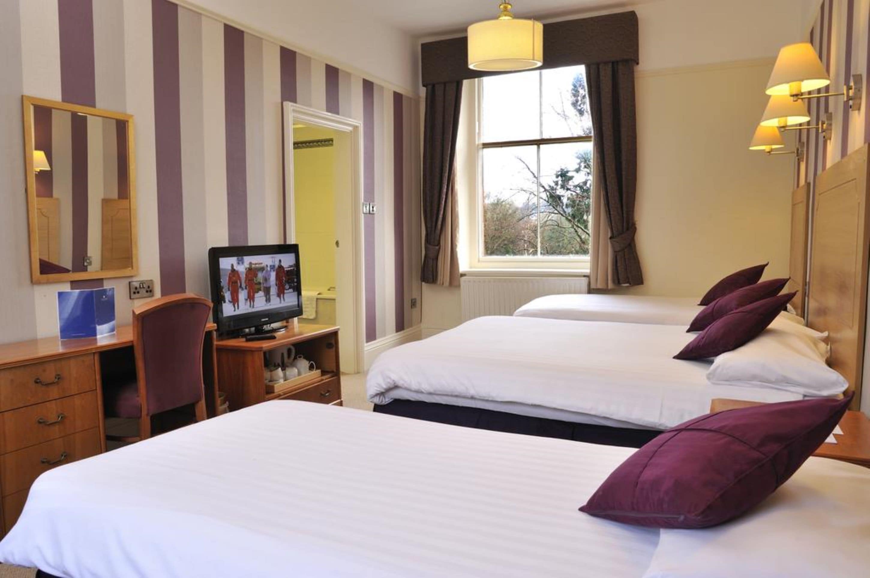 A hotel room with striped wallpaper, a desk with a computer, two beds with pillows, a television, and a window with curtains.