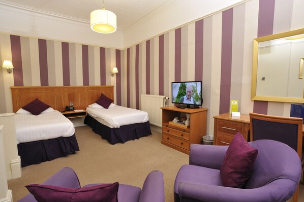 A hotel room with two single beds, a television, a wooden dresser, and a purple armchair.