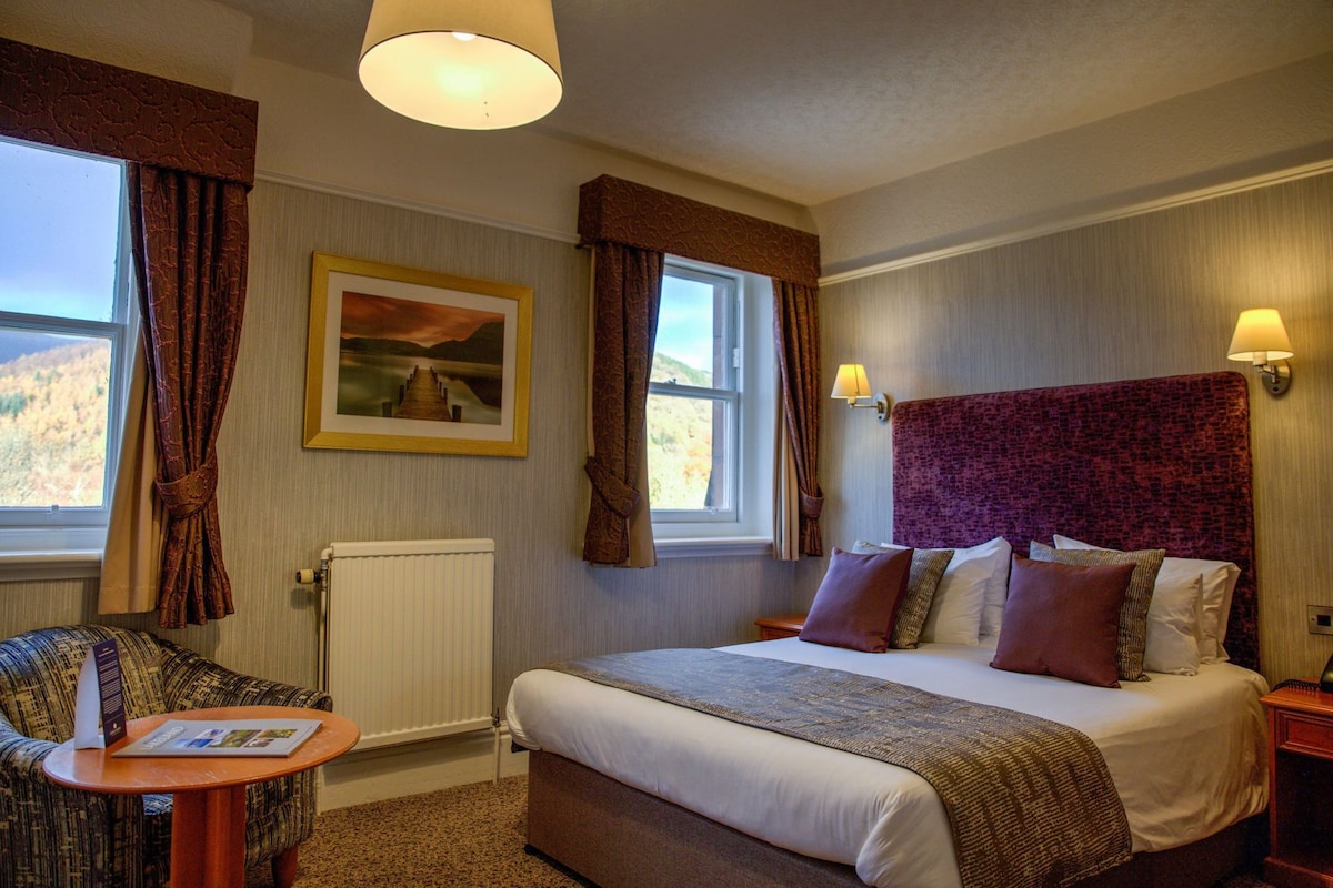 A hotel room with a large bed, a small table, a chair, a framed picture on the wall, and a window with curtains.