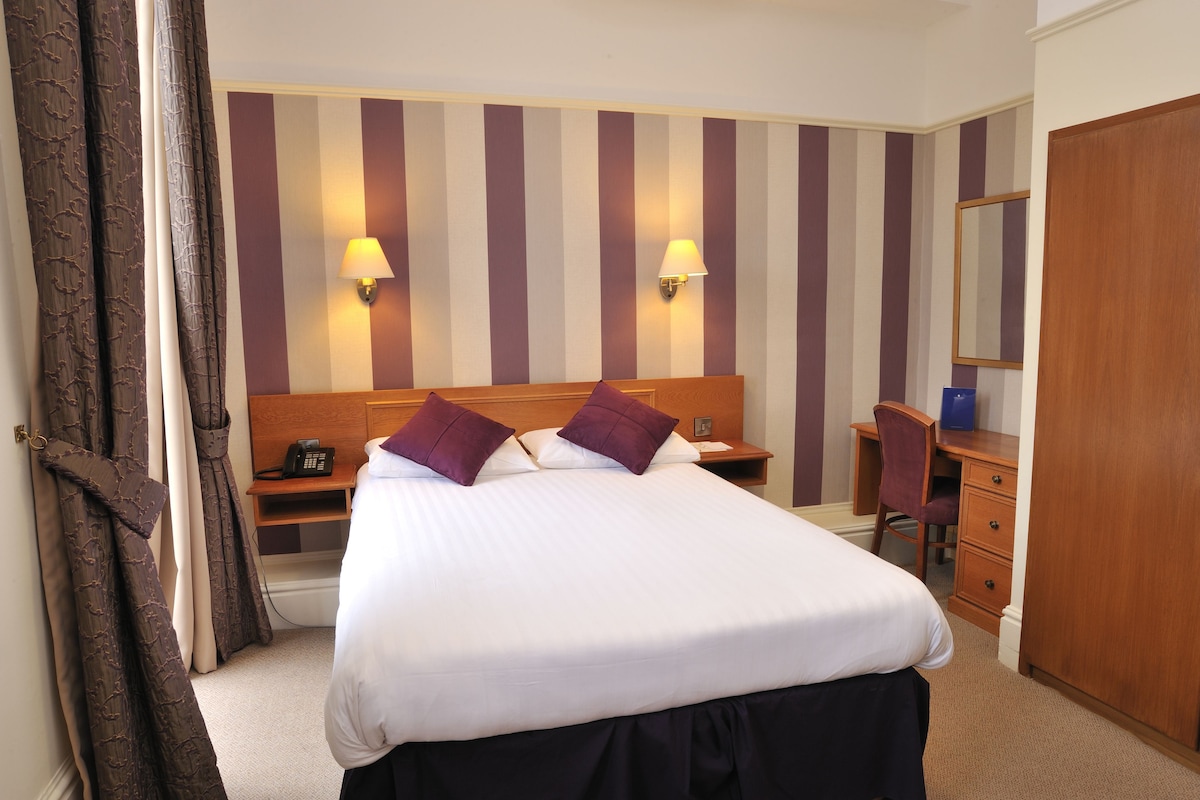 A hotel room with a bed, bedside lamps, a desk, a chair, a wardrobe, and a mirror.
