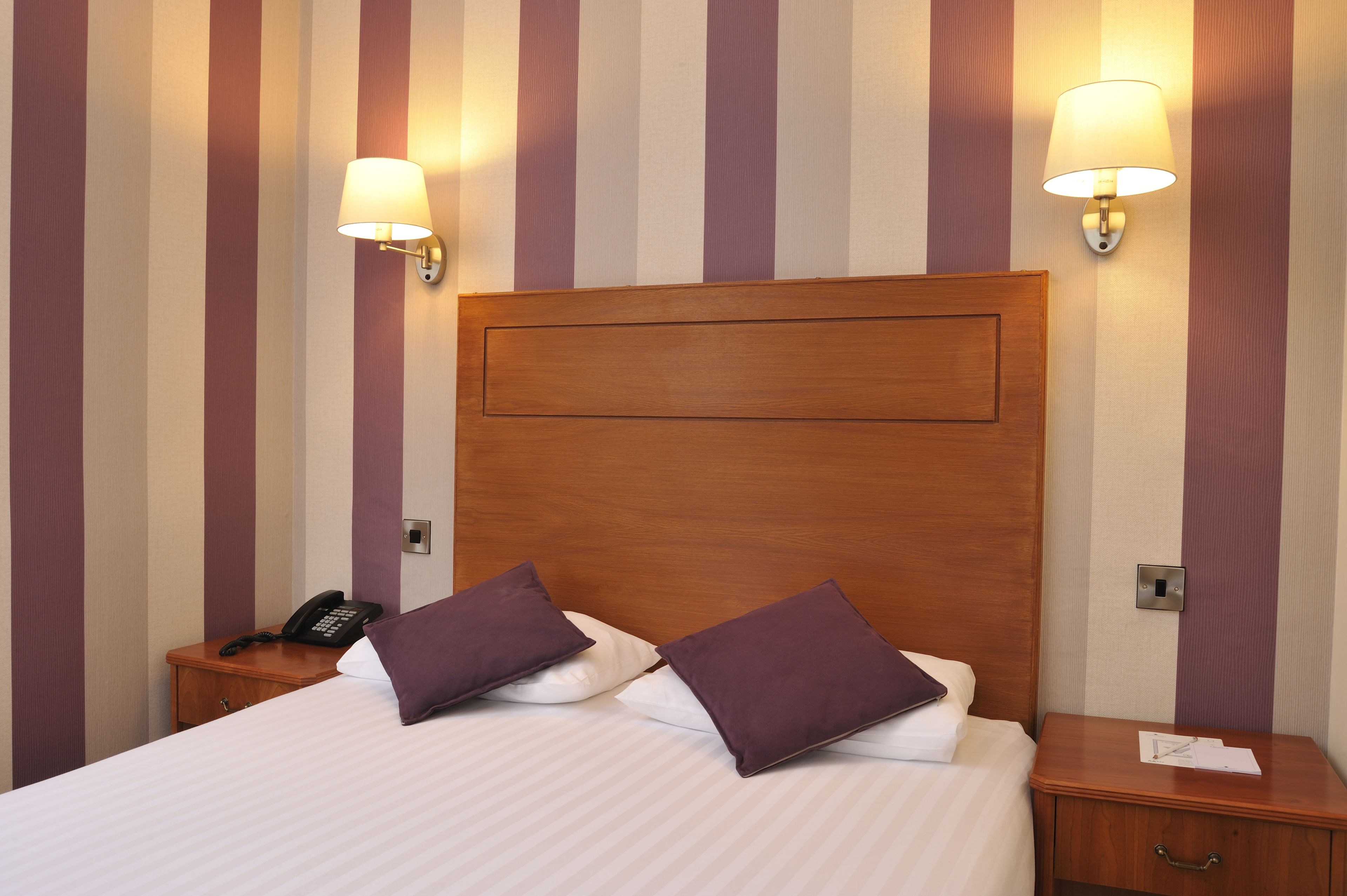 Habitación de hotel con una cama grande, dos mesitas de noche, un cabecero de madera y dos lámparas de pared.