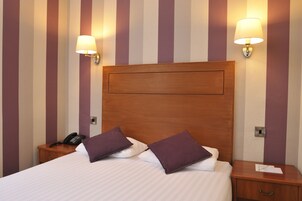 Habitación de hotel con una cama grande, dos mesitas de noche, un cabecero de madera y dos lámparas de pared.