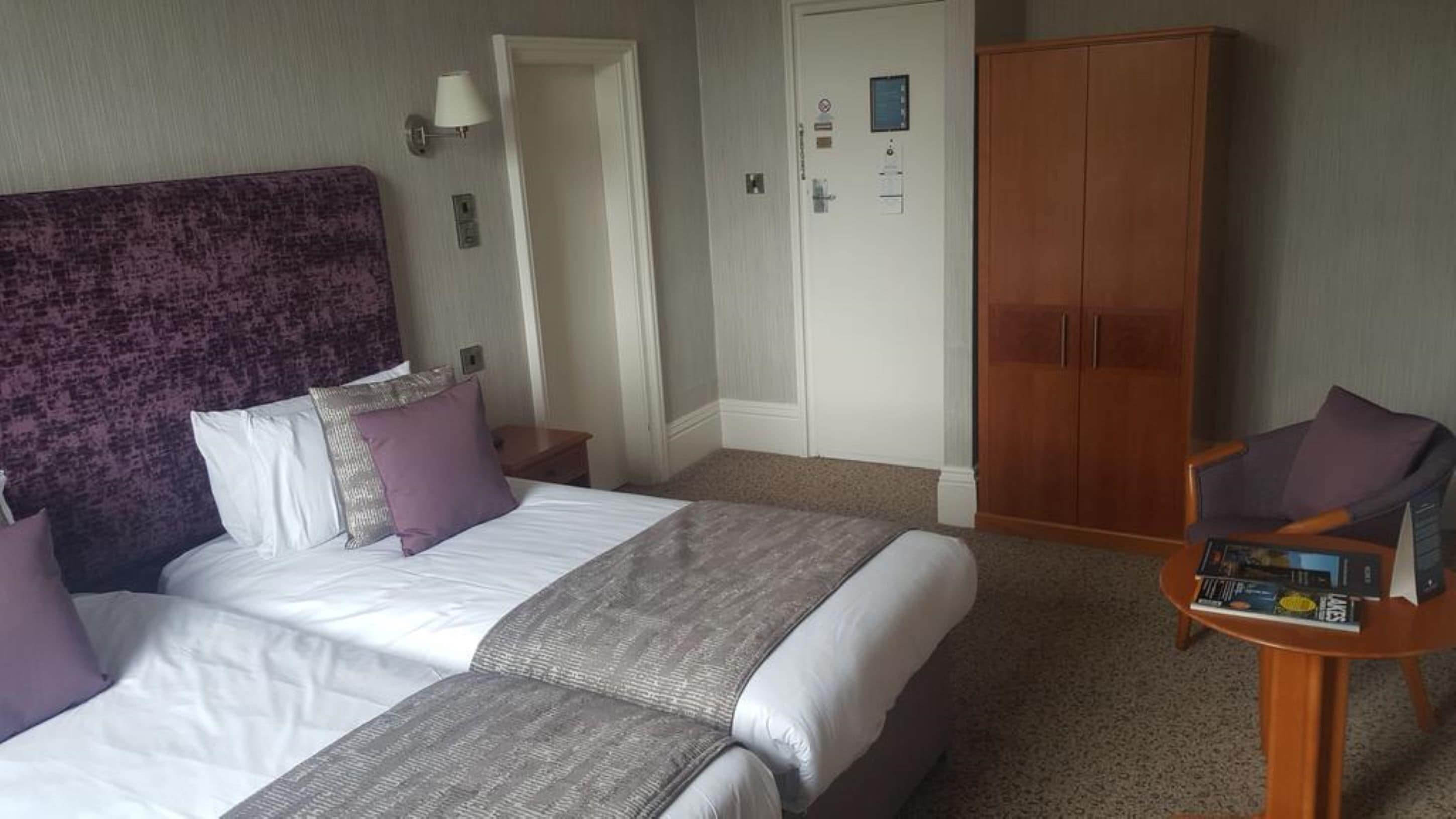 A hotel room with a large bed, a wooden wardrobe, a nightstand with a lamp, and a small table with magazines.