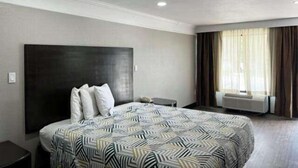 A hotel room with a large bed, a dark headboard, a bedside table, and a window with curtains. - Studio 6 Suites Hinesville, GA  (Hinesville)