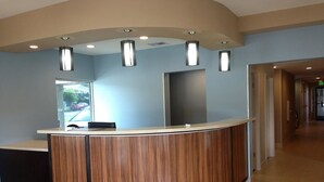 A reception desk with a curved design, a wooden finish, and a modern light fixture above it. - Studio 6 Suites Hinesville, GA  (Hinesville)