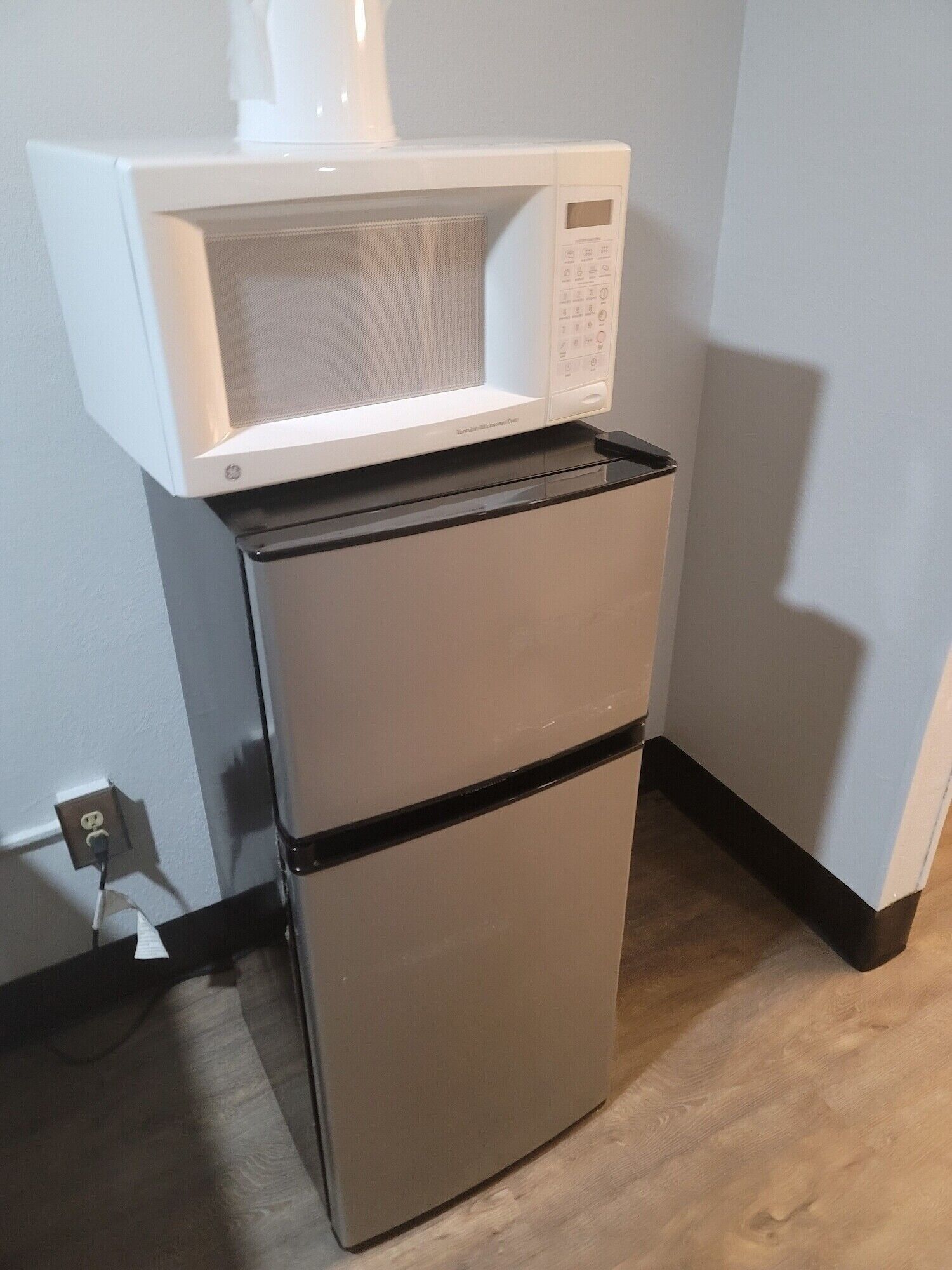 A stainless steel refrigerator with a microwave on top, located in a corner of a room with a wooden floor and a white wall.