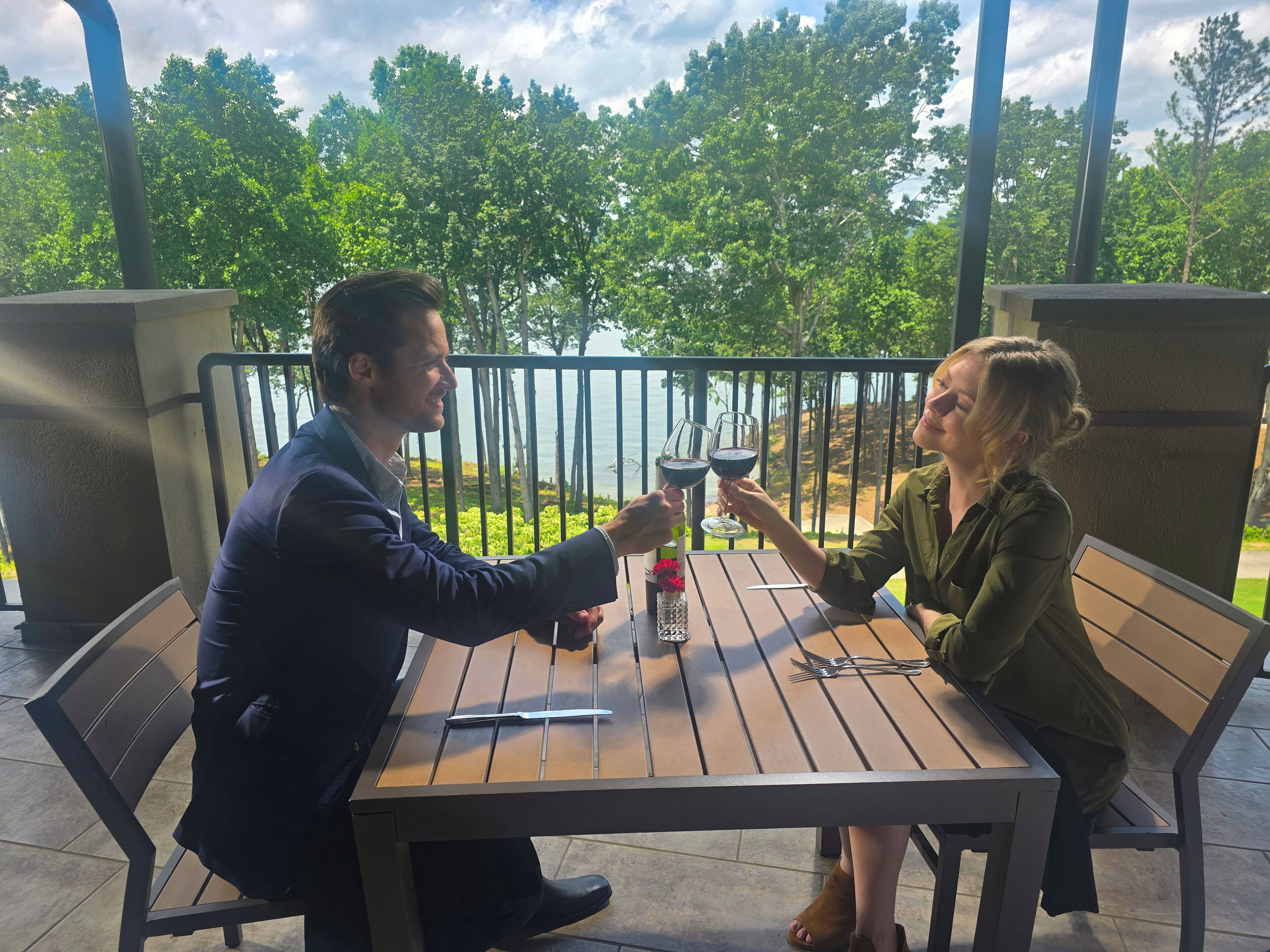 A man and a woman sitting at a wooden table on an outdoor patio with a view of a lake and trees.