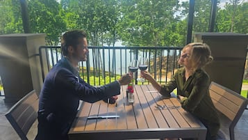 A man and a woman sitting at a wooden table on an outdoor patio with a view of a lake and trees.