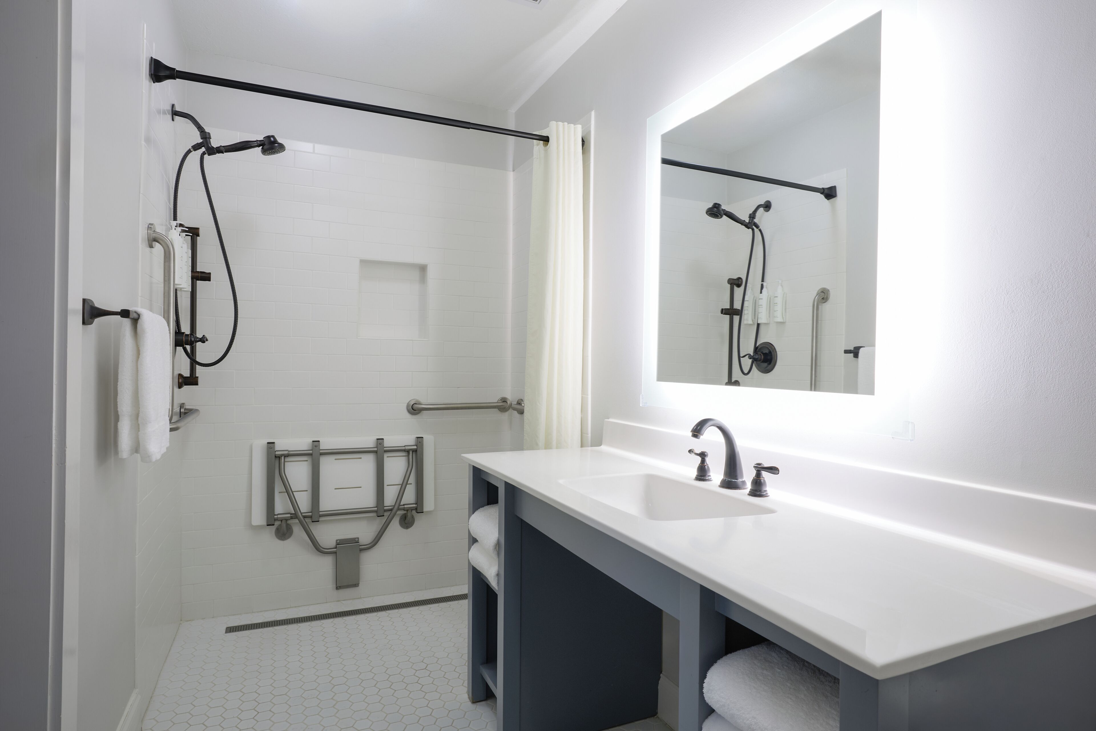 Un baño moderno con ducha a nivel, barras de apoyo, un espejo grande y un lavabo blanco.