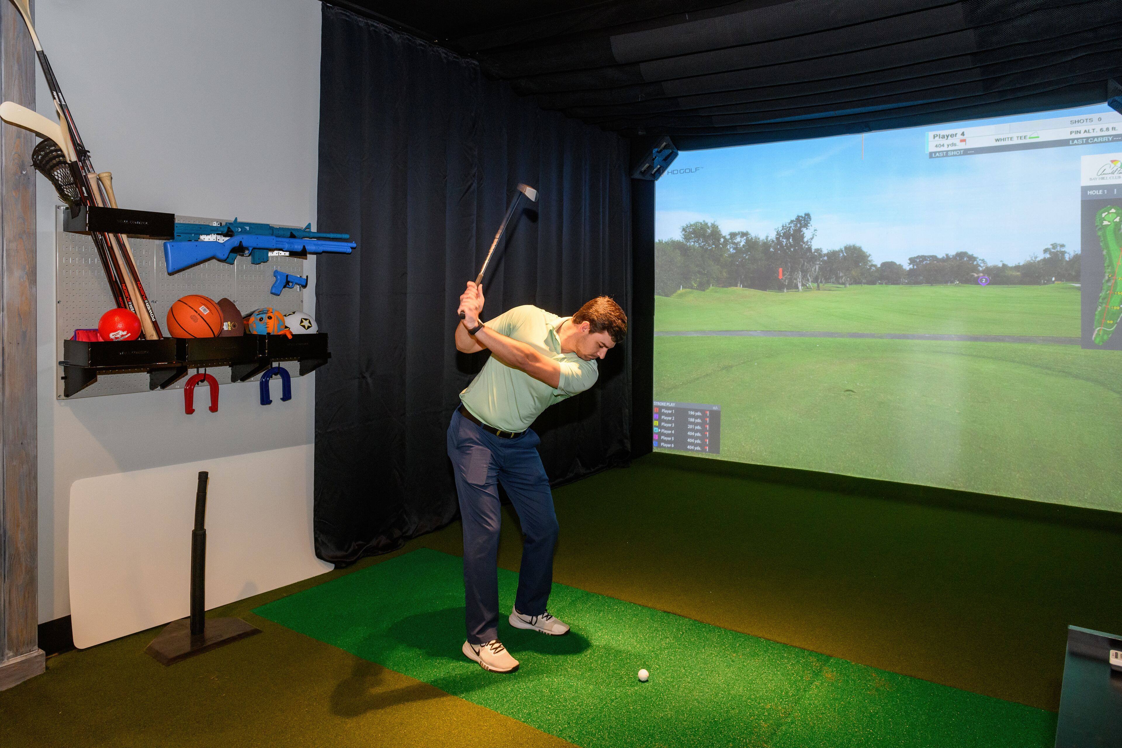 Ein Mann übt Golfschwünge in einem Raum mit grünem Turf-Boden, einem Bildschirm, der einen Golfplatz zeigt, und verschiedenen Sportgeräten an der Wand.