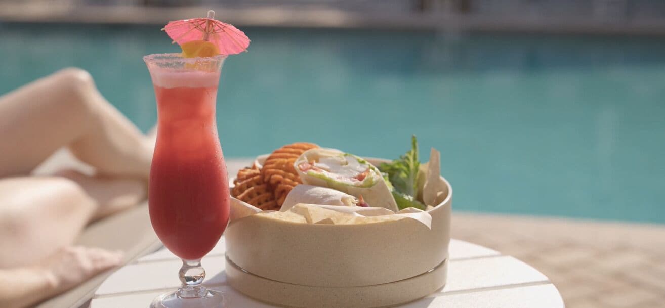 Een cocktail met een roze parasol, een saladeschaal en een bord crackers bij een zwembad.