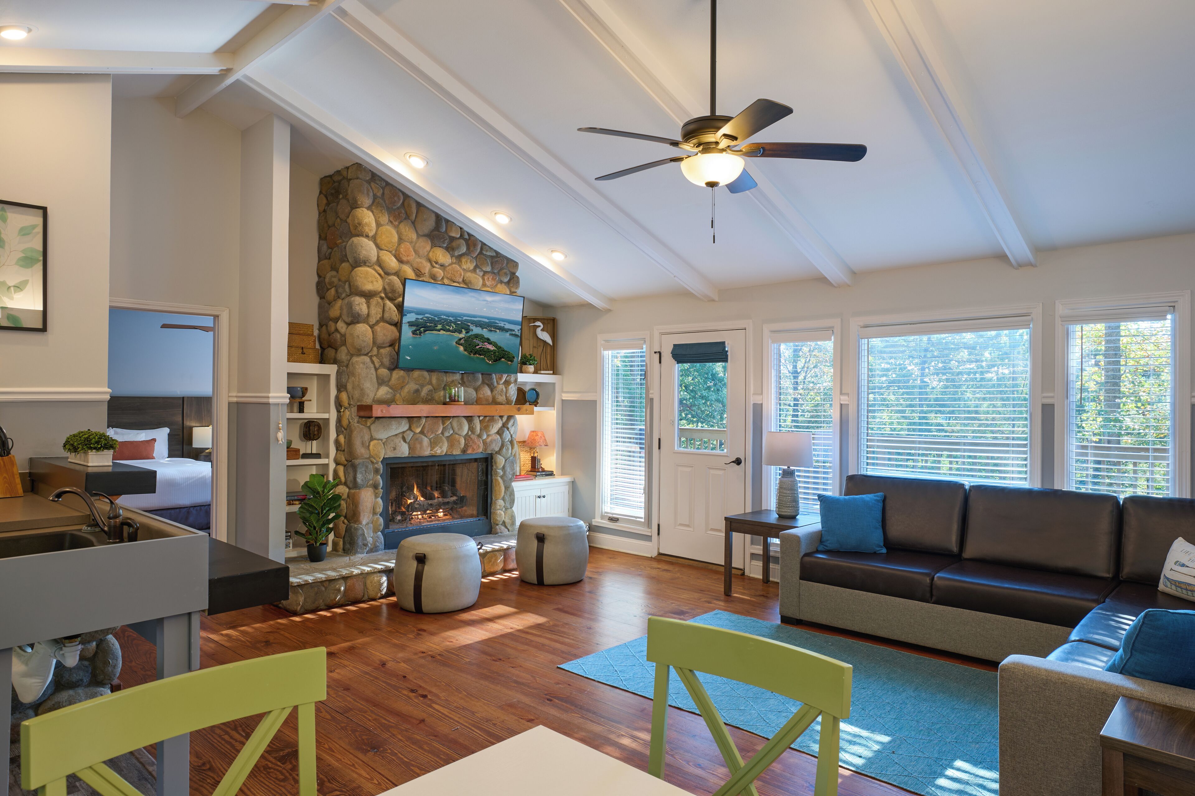 A spacious living room with a stone fireplace, a ceiling fan, and a large window with blinds.