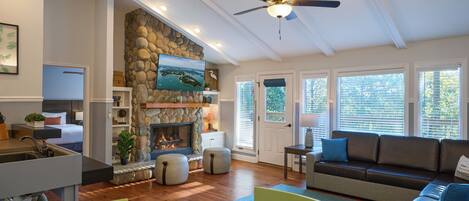 A spacious living room with a stone fireplace, a ceiling fan, and a large window with blinds.