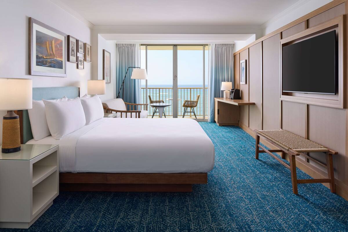 A hotel room with a large bed, a desk, a chair, a TV, and a balcony with a view.