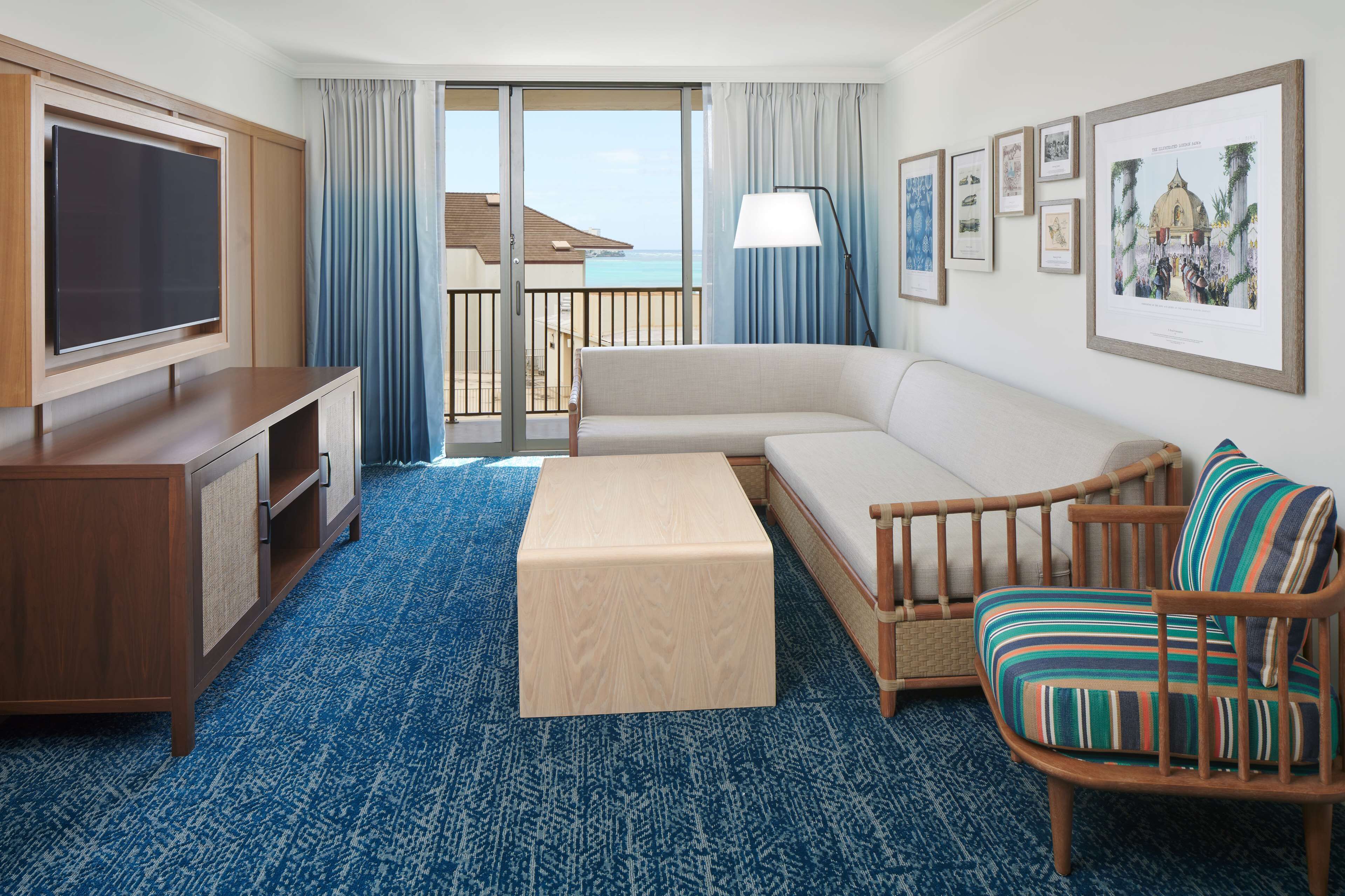 A hotel room with a large flat-screen TV, a sofa, a wooden chair, a small table, and a balcony with a view of the sea.