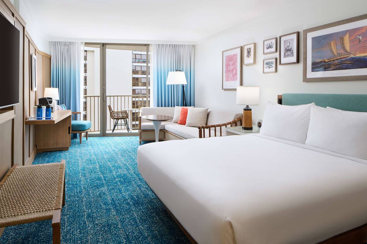 A hotel room with a large bed, a desk with a computer, a chair, a small table, and a balcony with curtains.