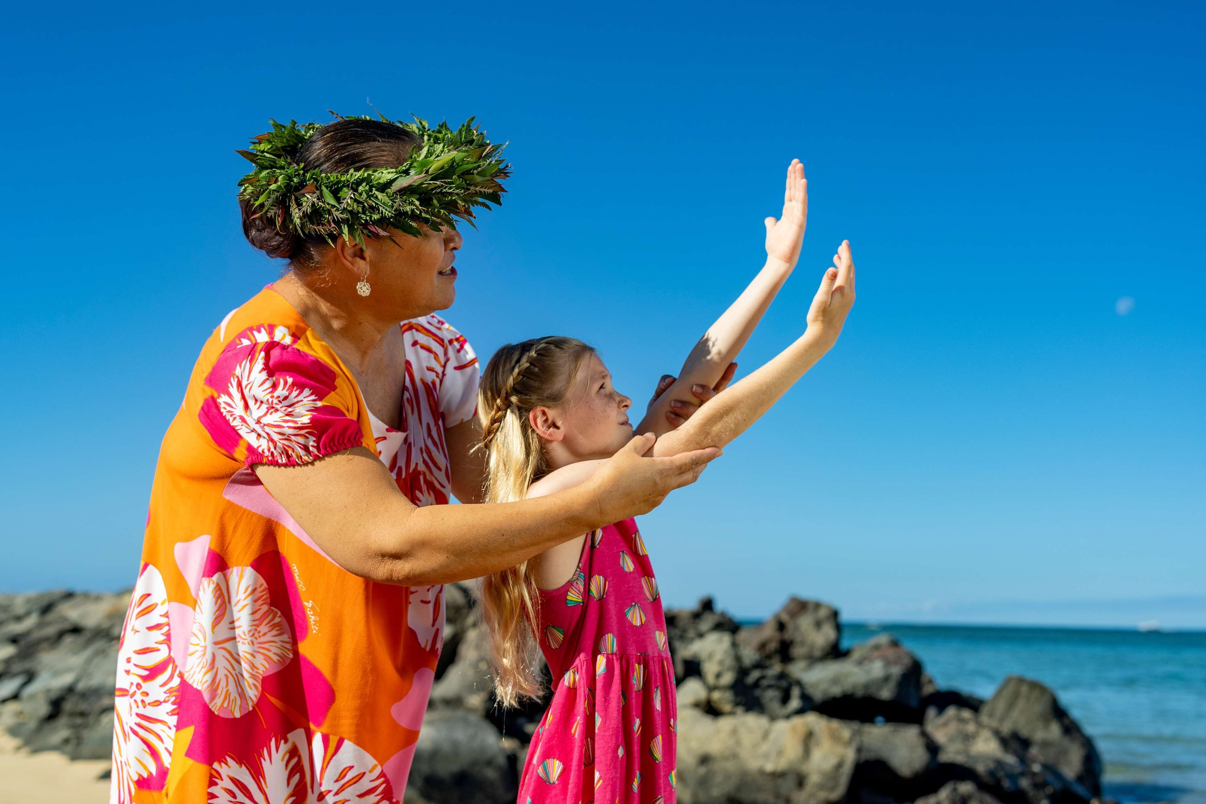 A person in a floral dress and a lei on their head, standing on a beach with rocks and water in the background.