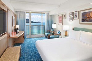 A hotel room with a bed, a desk, a chair, a small table, a lamp, a picture frame, and a view of the ocean. - OUTRIGGER Reef Waikiki Beach Resort (Honolulu)