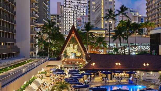 Un hotel con terraza exterior, piscina y un edificio destacado con estructura de techo triangular.
