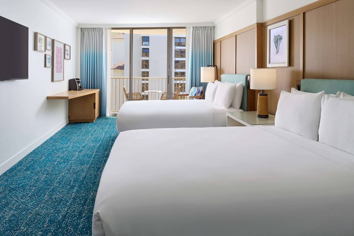 A hotel room with two beds, a desk, a chair, a TV, and a balcony with a view of buildings.
