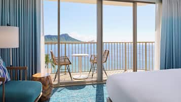 A hotel room with a large balcony, a bed, a chair, a table, and a view of the sea and a mountain.