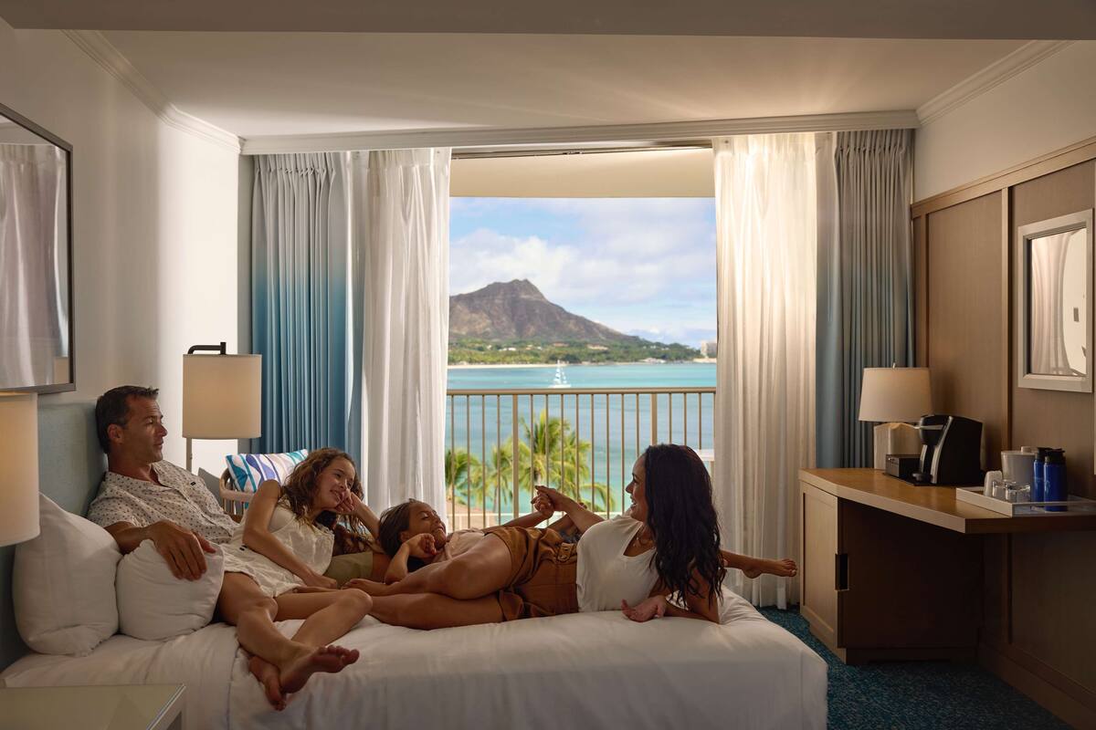 A hotel room with a bed, a desk, and a view of a beach and mountain.