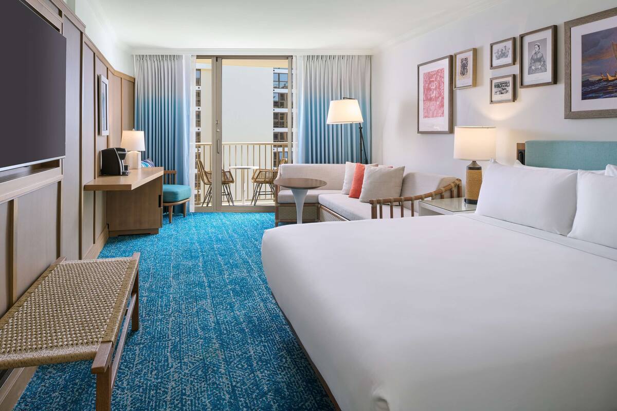 A hotel room with a large bed, a desk with a chair, a small table, a sofa, and a balcony with curtains.