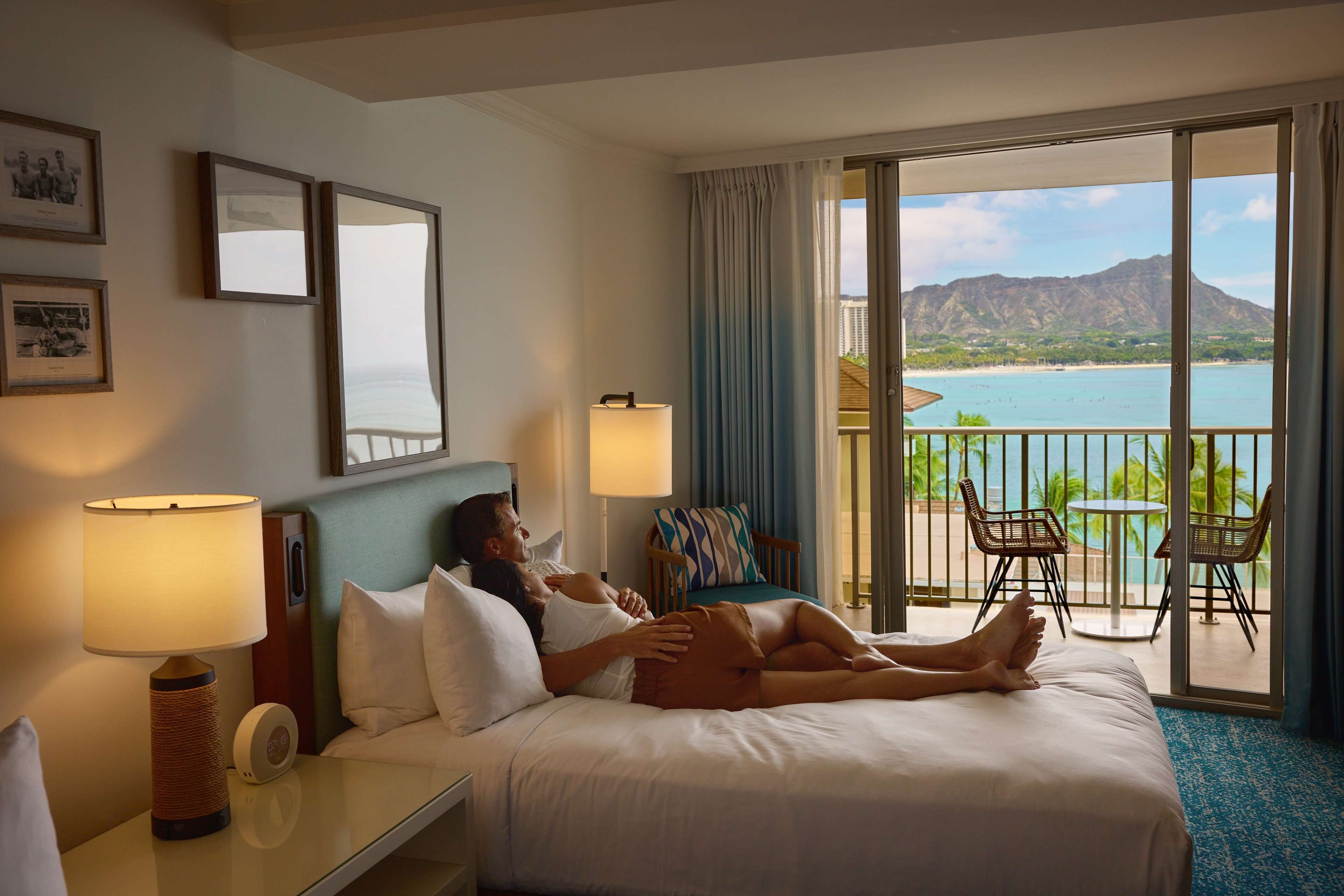 A hotel room with a bed, two bedside lamps, a nightstand, a chair, and a view of a beach and mountains.