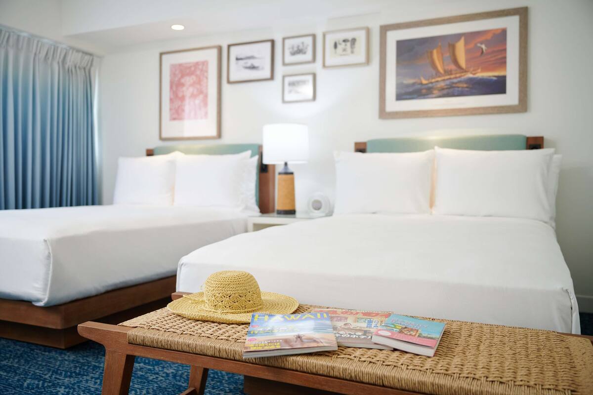 A hotel room with two beds, a wicker table with magazines, and framed artwork on the wall.