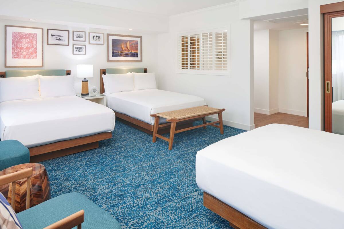 A hotel room with two beds, a small wooden bench, a nightstand with a lamp, and framed pictures on the wall.