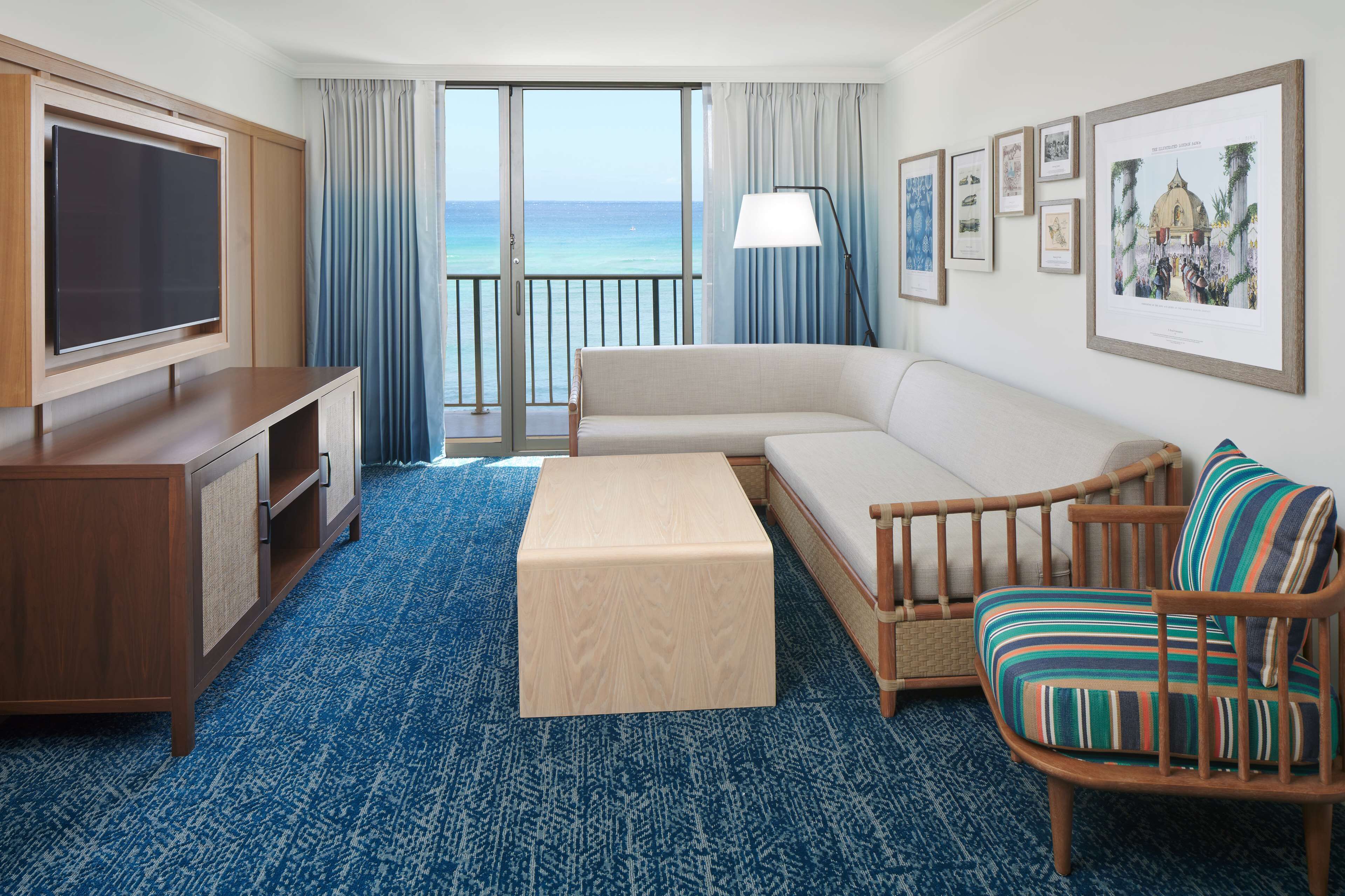 A hotel room with a large TV, a sofa, a wooden chair, a small table, and a balcony with a view of the ocean.