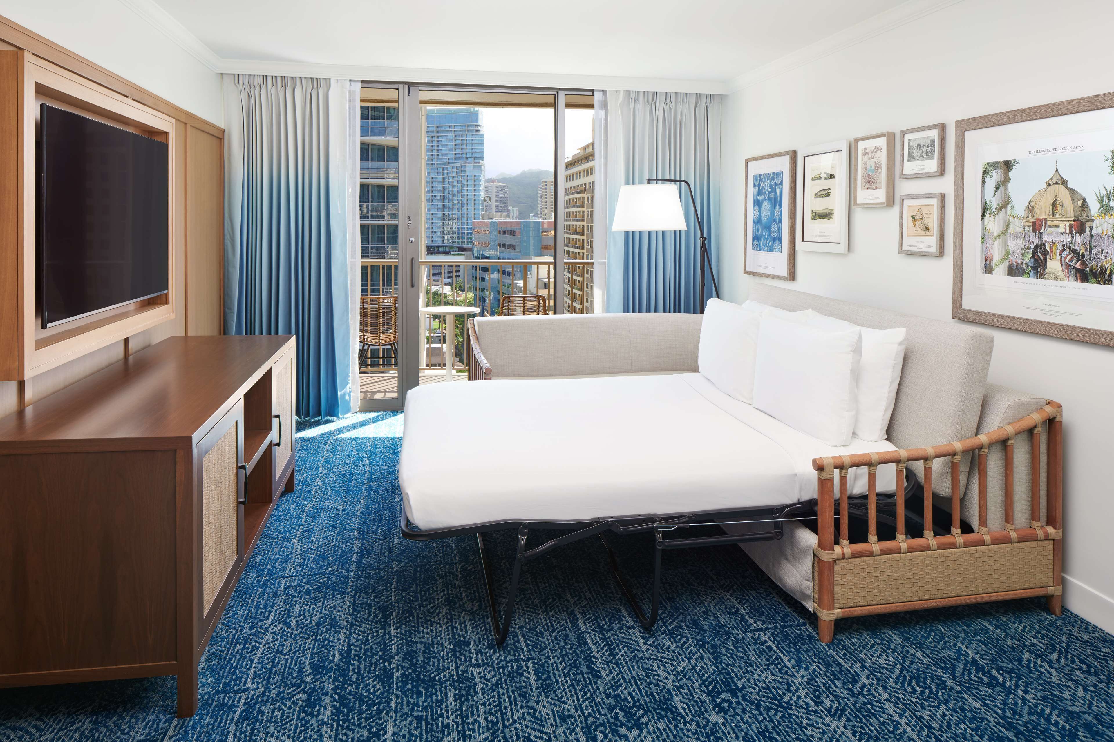 A hotel room with a bed, a TV, a desk, a balcony with a view of buildings, and framed pictures on the wall.