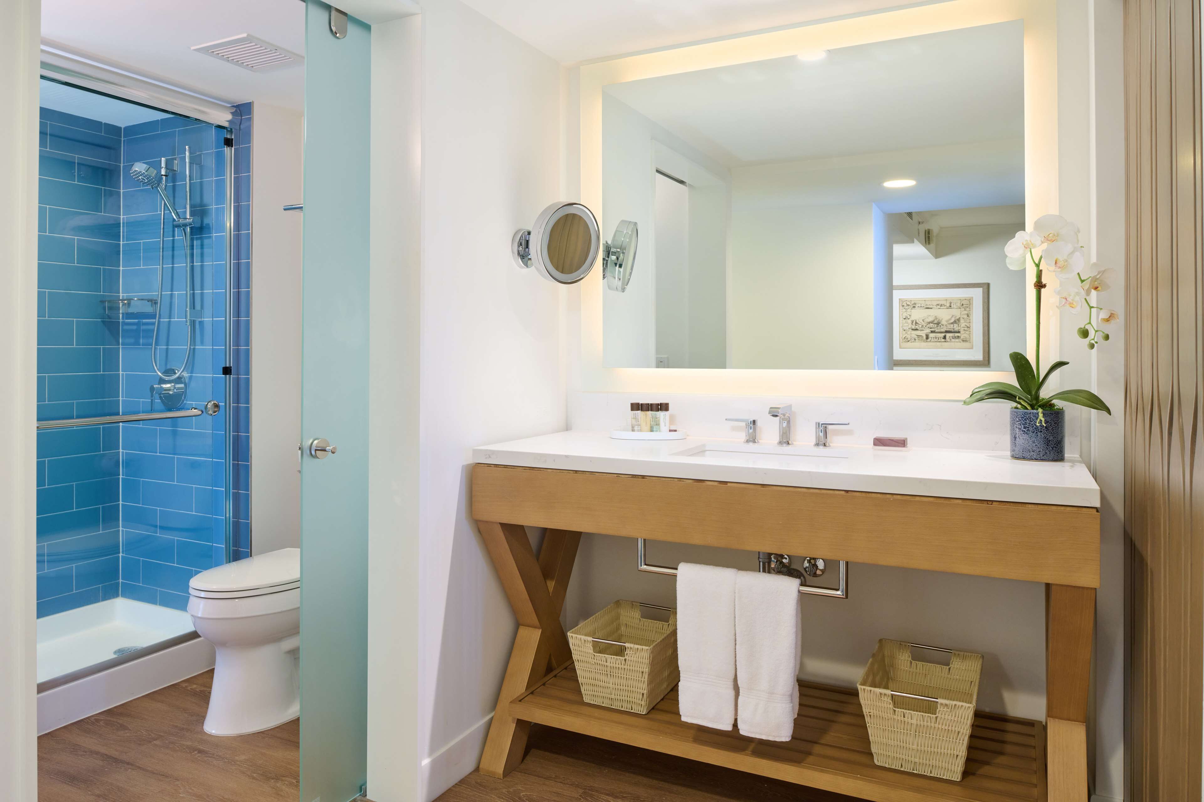 A bathroom with a glass-enclosed shower, a white toilet, a wooden vanity with a mirror, and a potted orchid.
