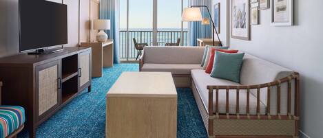 A hotel room with a large flat-screen TV, a sofa, a small wooden table, a baby crib, and a balcony with a view.