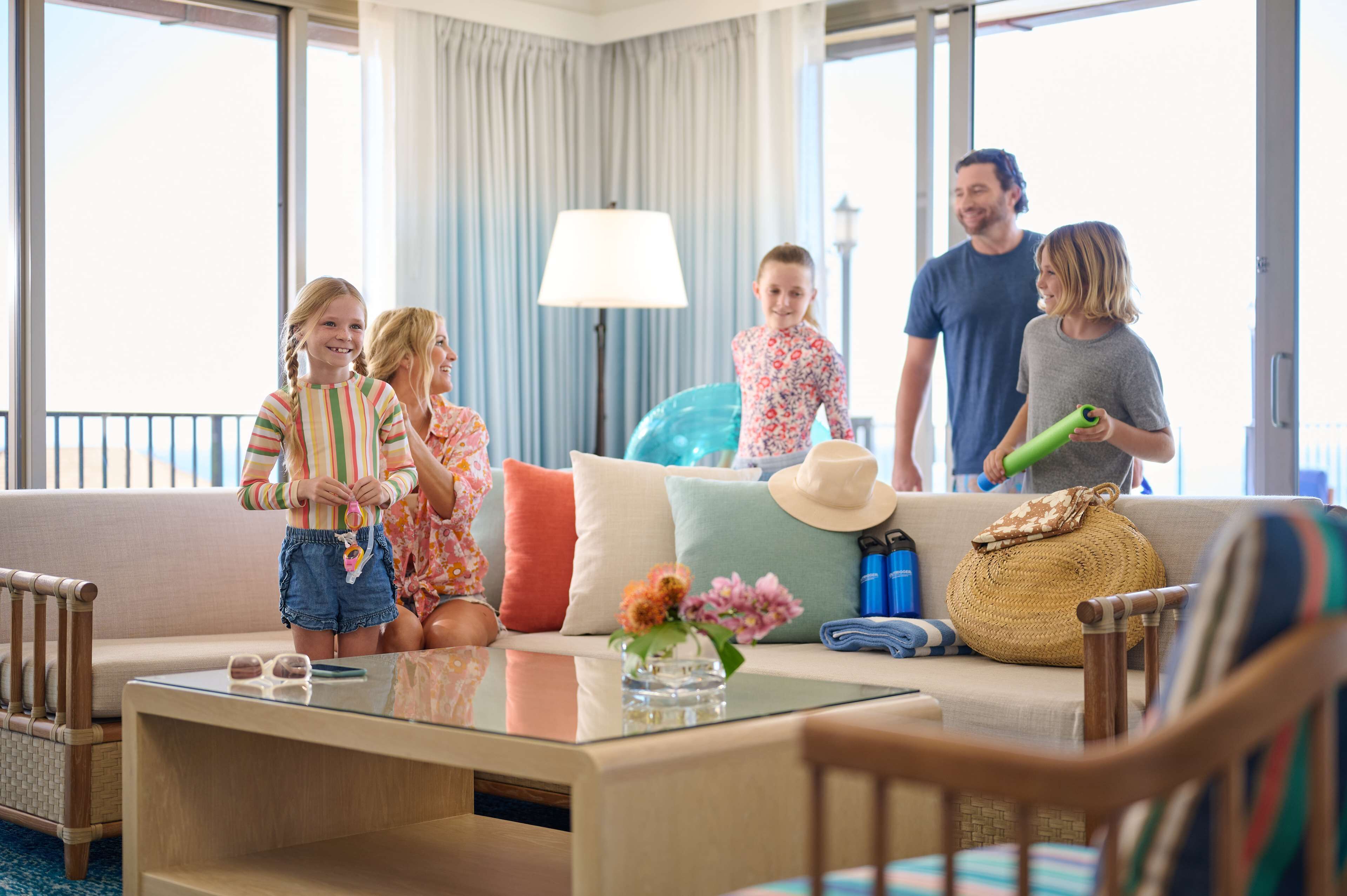 Eine Familie in einem Hotelzimmer mit einer Couch, einem Glastisch und Blick durch große Fenster.