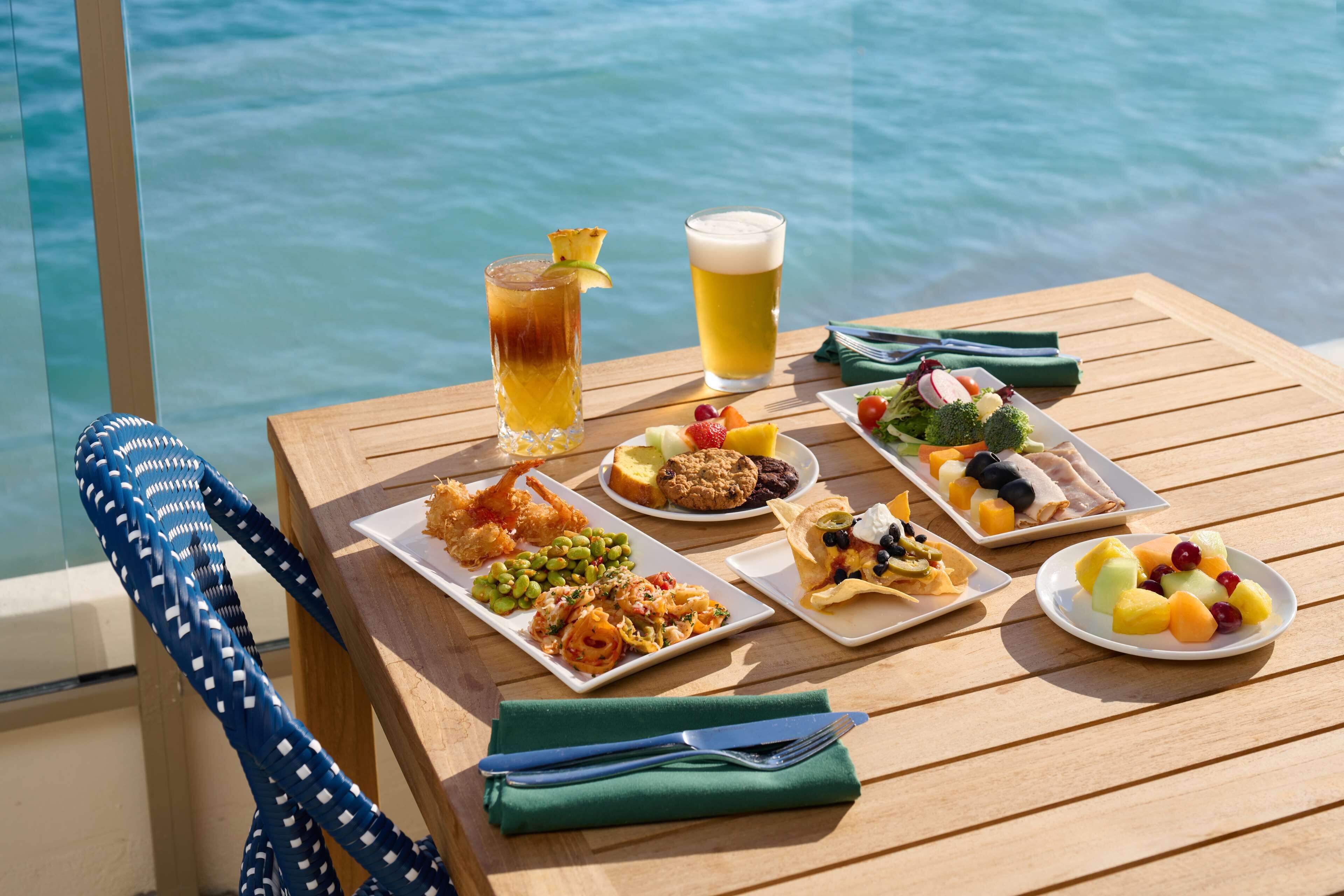 Uma mesa de madeira com diversos pratos e bebidas, com vista para um corpo de água.