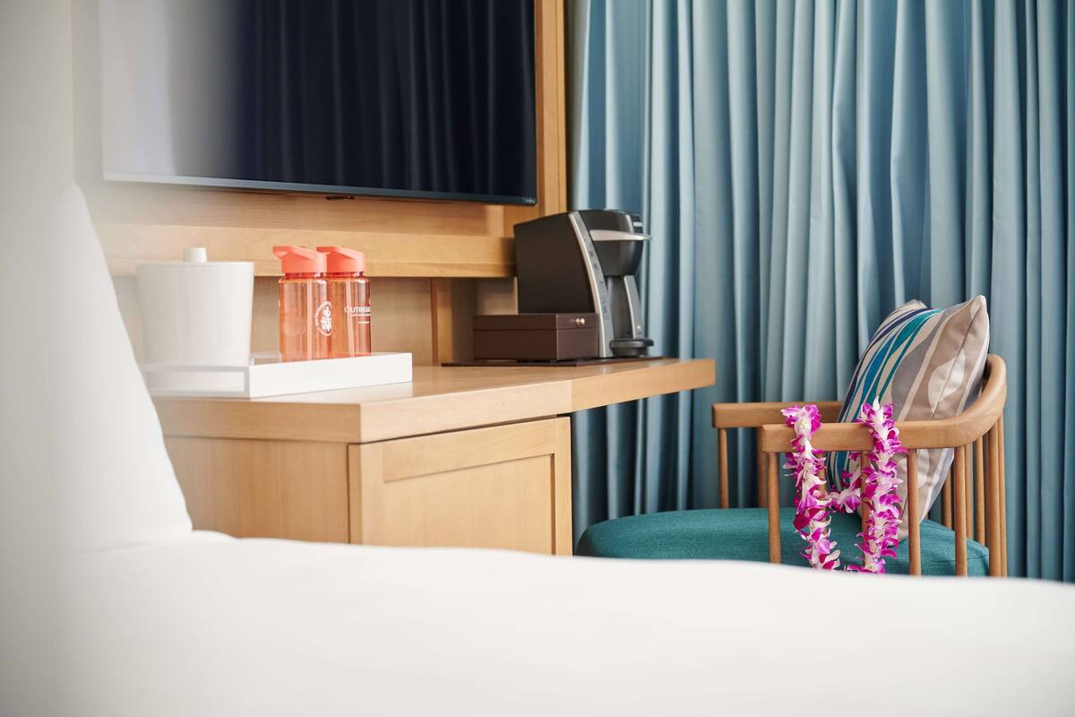 A hotel room with a bed, a desk with a coffee maker, a television, and a chair with a lei.