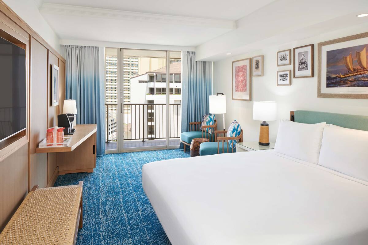 A hotel room with a large bed, a desk with a lamp, a chair, a small table with a lamp, and a view of a building through a glass door.