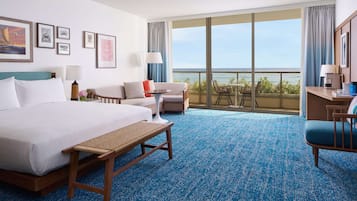 A hotel room with a large bed, a sofa, a small table, a bench, and a balcony with a view of the ocean.