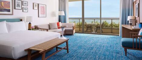Habitación de hotel con una cama grande, un sofá, una mesita, un banco y un balcón con vista al océano.