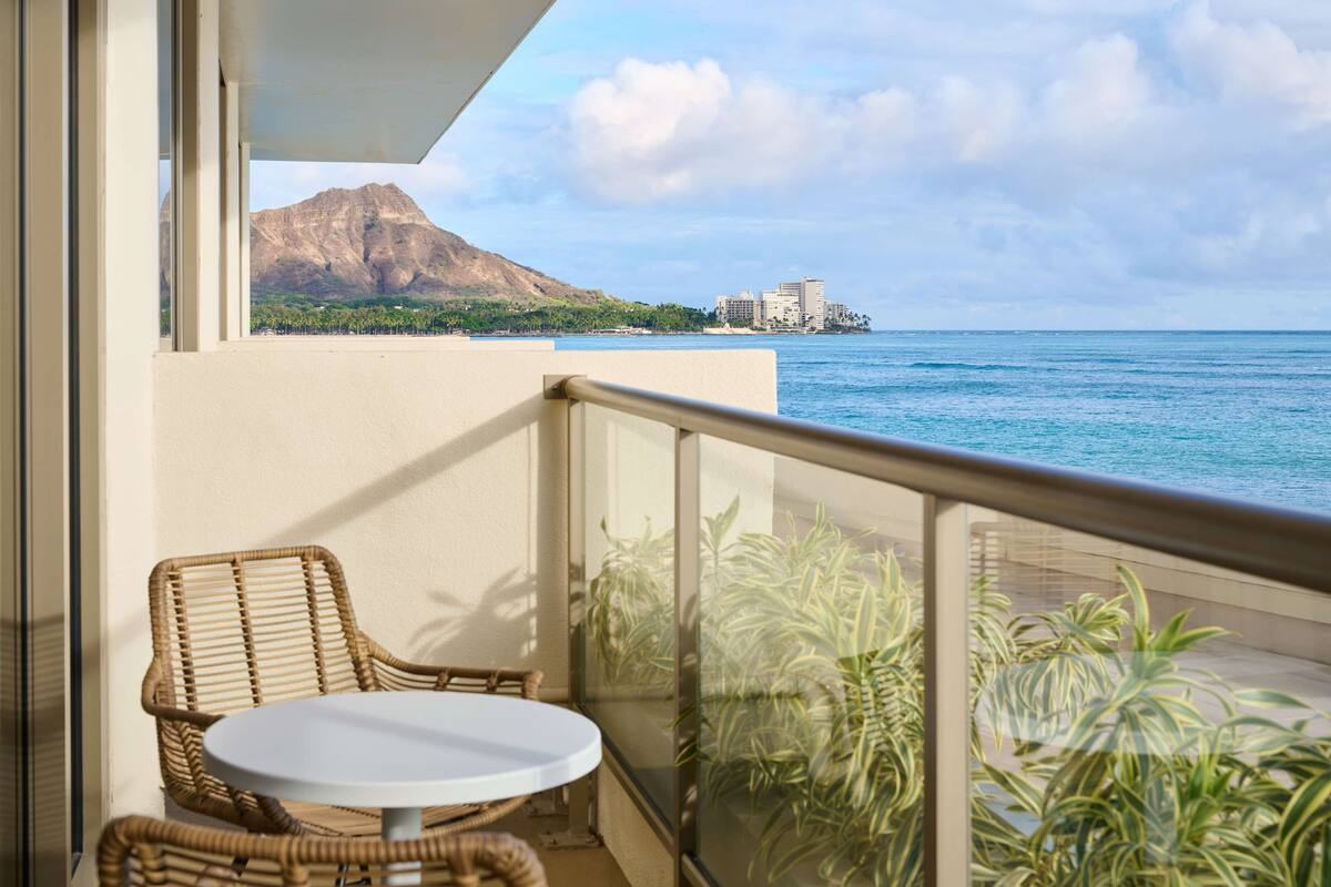 A balcony with wicker furniture, a glass railing, and a view of the ocean and a mountain.