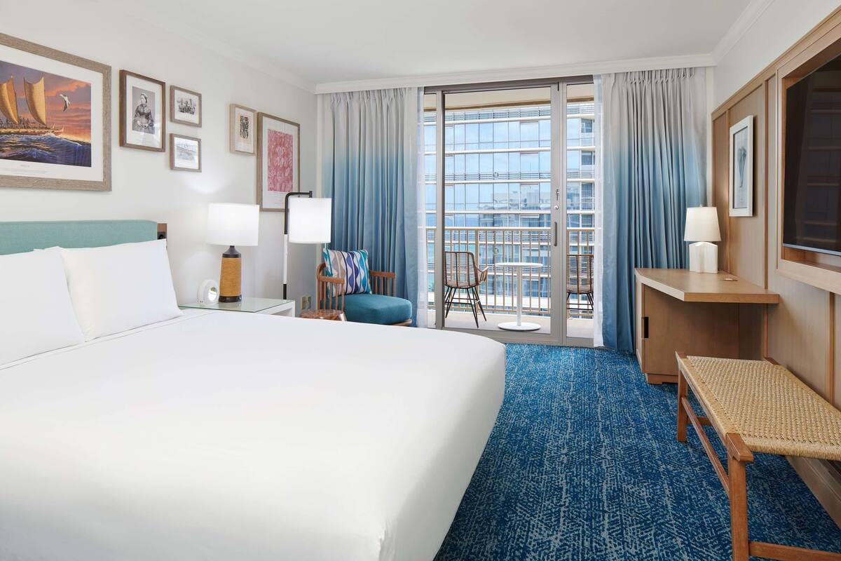 A hotel room with a large bed, a desk, a chair, a balcony with a view, and framed pictures on the wall.