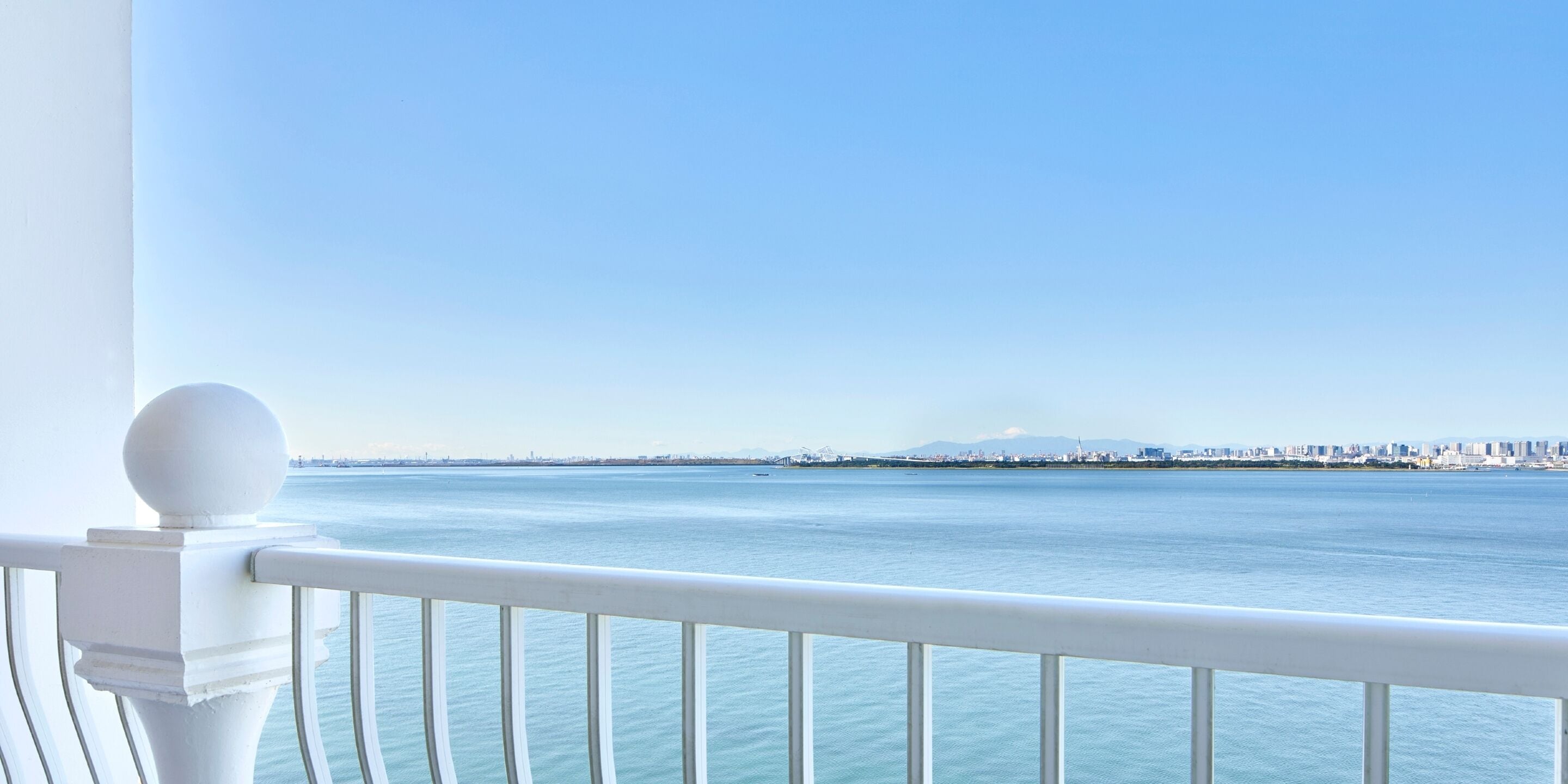 Un balcon doté d’une balustrade blanche, offrant une vue sur une étendue d’eau paisible et sur les contours d’une ville lointaine.